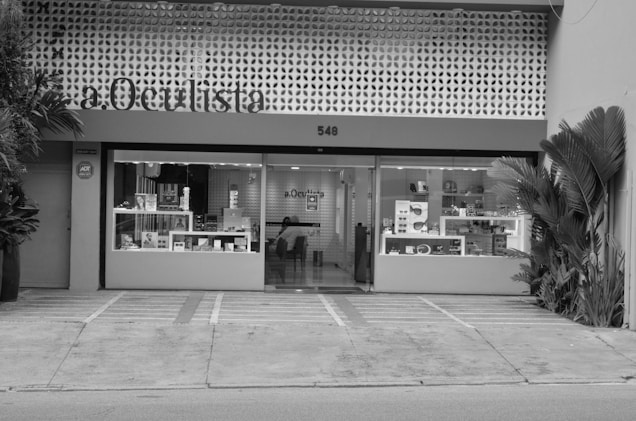 A building facade with a modern design, featuring a sign labeled 'a.Oculista'. The store has large windows displaying various items inside. Potted plants are positioned on both sides of the entrance. The exterior is characterized by a perforated pattern above the windows, and there appears to be a seating area with a person inside.