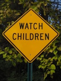 A diamond-shaped yellow traffic sign with the words 'WATCH CHILDREN' in bold black letters is prominently displayed. The background consists of dense green foliage and trees, suggesting the sign is located in a natural or suburban area.