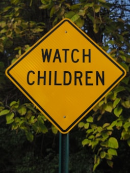 A diamond-shaped yellow traffic sign with the words 'WATCH CHILDREN' in bold black letters is prominently displayed. The background consists of dense green foliage and trees, suggesting the sign is located in a natural or suburban area.