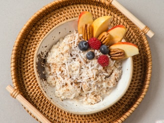 A bowl of oatmeal topped with shredded coconut, sliced apples, pecans, blueberries, and a raspberry, served on a woven tray.