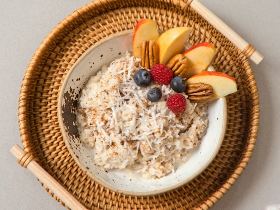 A bowl of oatmeal topped with shredded coconut, sliced apples, pecans, blueberries, and a raspberry, served on a woven tray.