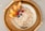 A bowl of oatmeal topped with shredded coconut, apple slices, pecans, blueberries, and raspberries, placed on a wicker tray with a simple handle design and a light gray background.