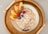 A bowl of oatmeal topped with shredded coconut, apple slices, pecans, blueberries, and raspberries, placed on a wicker tray with a simple handle design and a light gray background.