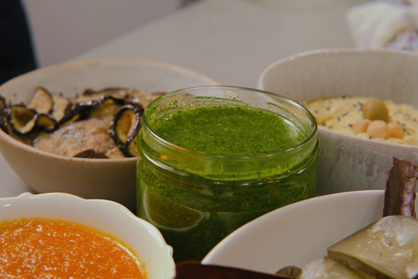 Several bowls and dishes containing various foods, including a vibrant green sauce or dip in a glass jar, an orange-colored sauce, sliced mushrooms on another dish, and a dish with chickpeas.