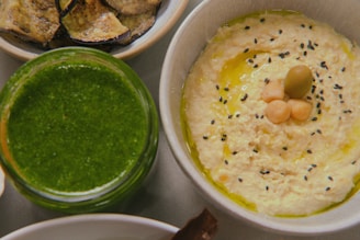 A close-up of a bowl of hummus garnished with black sesame seeds, chickpeas, and a green olive, arranged in the center. Next to it, there is a jar filled with a bright green pesto-like sauce. In the background, a plate with roasted eggplant slices is partially visible.