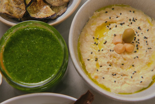 A close-up of a bowl of hummus garnished with black sesame seeds, chickpeas, and a green olive, arranged in the center. Next to it, there is a jar filled with a bright green pesto-like sauce. In the background, a plate with roasted eggplant slices is partially visible.