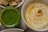 A close-up of a bowl of hummus garnished with black sesame seeds, chickpeas, and a green olive, arranged in the center. Next to it, there is a jar filled with a bright green pesto-like sauce. In the background, a plate with roasted eggplant slices is partially visible.