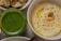 A close-up of a bowl of hummus garnished with black sesame seeds, chickpeas, and a green olive, arranged in the center. Next to it, there is a jar filled with a bright green pesto-like sauce. In the background, a plate with roasted eggplant slices is partially visible.