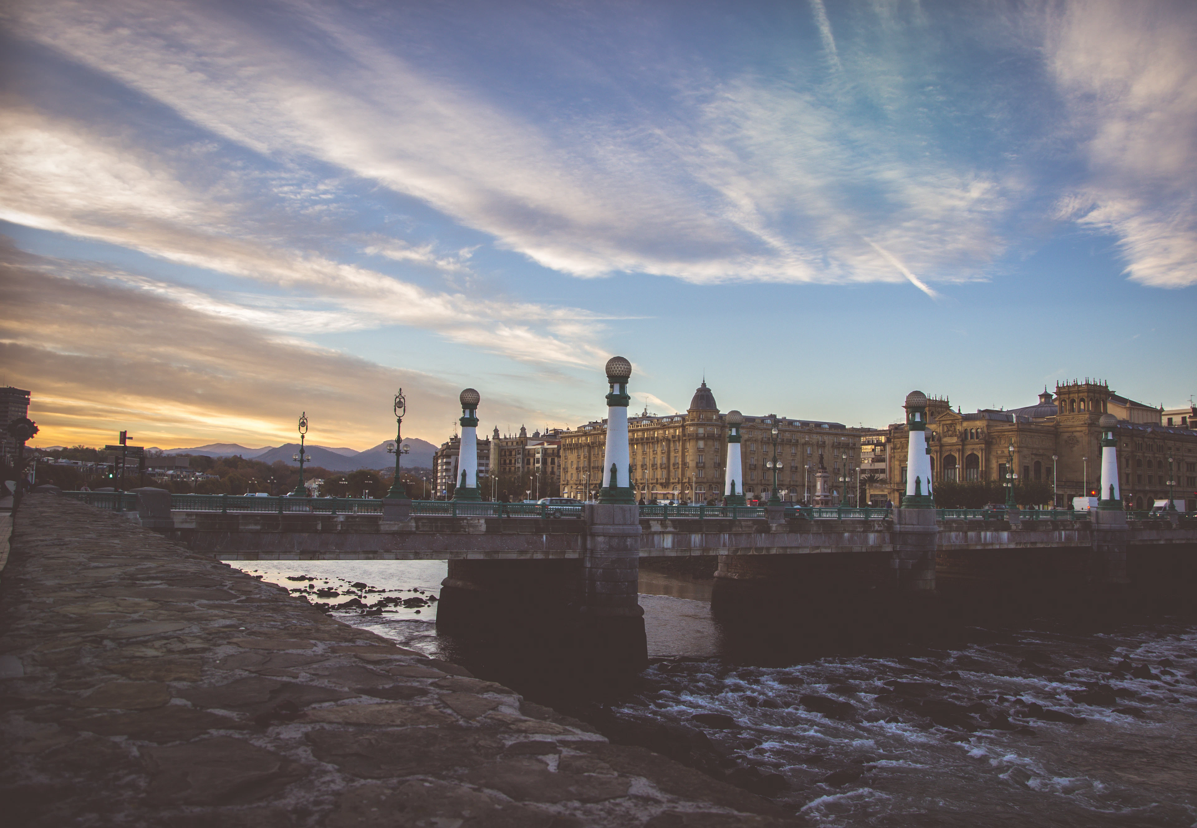 Sunset over the Urumea river in San Sebastian, a stone road bridge lined with tall white lamp columns crossing towards the belle-epoque buildings of the city, with the Basque hills dark on the horizon