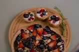 A wooden platter displaying a chocolate-covered dessert topped with sliced strawberries, blueberries, and almond pieces. Accompanying the dessert are three small tarts, each filled with yogurt and garnished with blackberries, blueberries, and red fruit pieces, along with a sprig of rosemary.