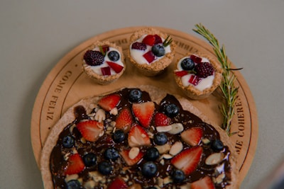 A wooden platter displaying a chocolate-covered dessert topped with sliced strawberries, blueberries, and almond pieces. Accompanying the dessert are three small tarts, each filled with yogurt and garnished with blackberries, blueberries, and red fruit pieces, along with a sprig of rosemary.
