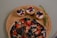A wooden platter displaying a chocolate-covered dessert topped with sliced strawberries, blueberries, and almond pieces. Accompanying the dessert are three small tarts, each filled with yogurt and garnished with blackberries, blueberries, and red fruit pieces, along with a sprig of rosemary.