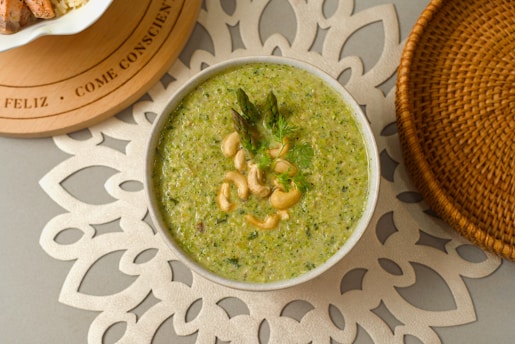 A bowl of green soup garnished with asparagus and a sprig of cilantro, with cashews on top, resting on an intricately designed white placemat. To the left, part of a circular wooden board with text can be seen, along with a wicker basket partially visible on the right.