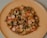 A homemade pizza topped with sliced mushrooms, sausages, bell peppers, and melted cheese is placed on a light wooden plate against a neutral background.