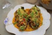 Two halves of vegetables, possibly stuffed or baked, are placed on a decorative plate with a patterned border. They are garnished with fresh green herbs and small red pieces, possibly chili or tomato, atop a base of orange sauce.