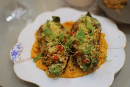 Two halves of vegetables, possibly stuffed or baked, are placed on a decorative plate with a patterned border. They are garnished with fresh green herbs and small red pieces, possibly chili or tomato, atop a base of orange sauce.