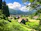 A tranquil rural scene featuring traditional thatched-roof houses nestled within lush greenery. Tall trees frame the image, and rolling hills stretch into the horizon under a bright blue sky filled with fluffy white clouds.