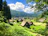 A tranquil rural scene featuring traditional thatched-roof houses nestled within lush greenery. Tall trees frame the image, and rolling hills stretch into the horizon under a bright blue sky filled with fluffy white clouds.