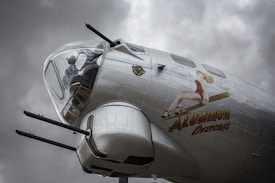 A vintage aircraft with a World War II-era nose art, featuring a pin-up girl sitting on a bomb, is prominently displayed against a backdrop of cloudy skies. The aircraft has a glossy metallic surface with visible panel lines and rivets.