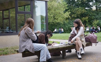 A group of three people are seated on wooden benches outside near a building. They are engaged in different activities, including writing and conversing. Backpacks are placed next to them. In the background, more people are seen sitting and interacting at picnic tables on a grassy area surrounded by trees.