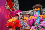 A vibrant display featuring colorful marigold and chrysanthemum flowers arranged around decorative elements, including a skeletal figurine dressed in traditional attire. The centerpiece includes a framed photograph of a man, suggesting a tribute or altar setup.