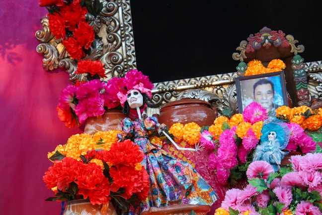 A vibrant display featuring colorful marigold and chrysanthemum flowers arranged around decorative elements, including a skeletal figurine dressed in traditional attire. The centerpiece includes a framed photograph of a man, suggesting a tribute or altar setup.