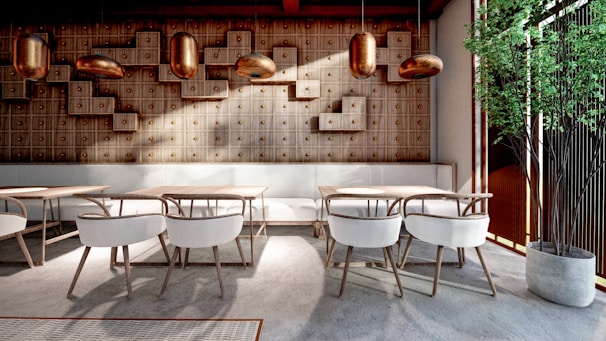 A modern, stylish interior with wooden walls featuring geometric patterns and warm lighting. The space includes sleek tables and chairs with light-colored upholstery, complemented by a potted green plant in the corner. The design is minimalistic yet inviting.