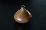 A ceramic vase with a rounded body and small narrow neck sits on a dark surface. The vase features a rich, earthy glaze with shades of brown and subtle hints of yellow and orange, giving it a textured and speckled appearance.
