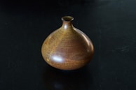 A ceramic vase with a rounded body and small narrow neck sits on a dark surface. The vase features a rich, earthy glaze with shades of brown and subtle hints of yellow and orange, giving it a textured and speckled appearance.