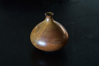 A ceramic vase with a rounded body and small narrow neck sits on a dark surface. The vase features a rich, earthy glaze with shades of brown and subtle hints of yellow and orange, giving it a textured and speckled appearance.