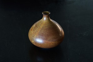 A ceramic vase with a rounded body and small narrow neck sits on a dark surface. The vase features a rich, earthy glaze with shades of brown and subtle hints of yellow and orange, giving it a textured and speckled appearance.