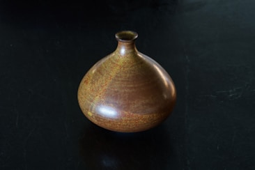 A ceramic vase with a rounded body and small narrow neck sits on a dark surface. The vase features a rich, earthy glaze with shades of brown and subtle hints of yellow and orange, giving it a textured and speckled appearance.