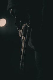 A dimly lit scene features a person wearing dark gloves holding a revolver. The lighting creates a dramatic and ominous atmosphere with deep shadows and minimal highlights.
