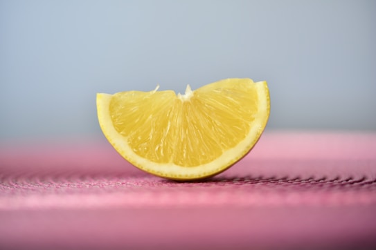 A slice of lemon stands upright on a textured pink surface with a soft, gradient background transitioning from blue to light gray. The lemon is fresh and juicy, with vibrant yellow rind and flesh.