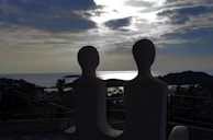 Two abstract sculptures are silhouetted against a serene coastal landscape. The sky is partly cloudy, and the sun is shining on the water, creating a reflective glow. There are hills and vegetation surrounding a body of water, likely a bay or harbor.