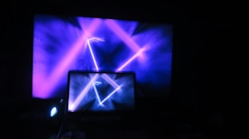 A laptop displaying vibrant, abstract blue and purple light patterns with a large screen in the background showing similar graphics. The scene is illuminated predominantly by electronic screens, creating a digital and futuristic atmosphere.