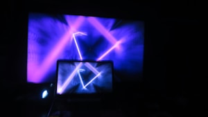 A laptop displaying vibrant, abstract blue and purple light patterns with a large screen in the background showing similar graphics. The scene is illuminated predominantly by electronic screens, creating a digital and futuristic atmosphere.