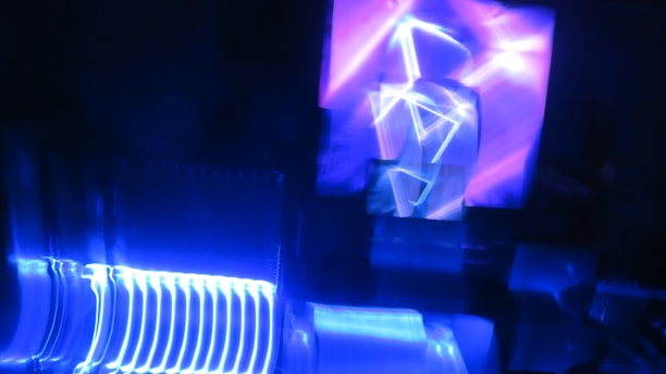 An abstract composition of neon lights creating geometric patterns against a dark background. The main colors are vivid blues with streaks of bright light forming angular shapes, giving a sense of movement and energy.