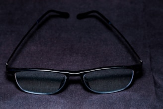A pair of rectangular glasses with dark frames is placed on a dark surface. The lenses appear clear and slightly reflective.