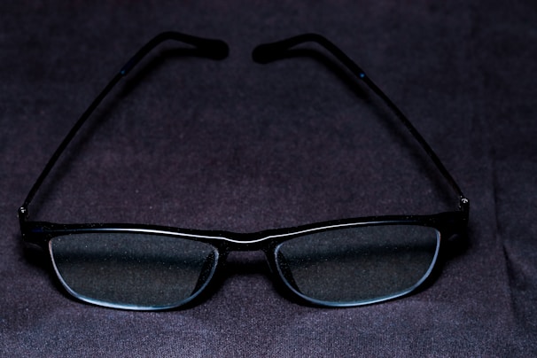 A pair of rectangular glasses with dark frames is placed on a dark surface. The lenses appear clear and slightly reflective.