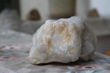 A large, rough stone with a primarily white surface and beige streaks sits on a table with a floral patterned cloth. The background is blurred, indicating a shallow depth of field, with indistinct shapes providing a neutral backdrop.
