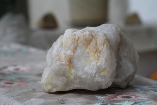A large, rough stone with a primarily white surface and beige streaks sits on a table with a floral patterned cloth. The background is blurred, indicating a shallow depth of field, with indistinct shapes providing a neutral backdrop.