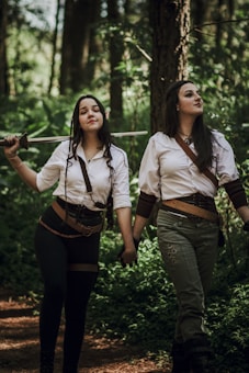 Two women dressed in medieval-inspired outfits walk hand in hand through a forest. They are both wearing white shirts and belts, with one holding a sword over her shoulder. The forest is lush and green, indicating a dense woodland environment.