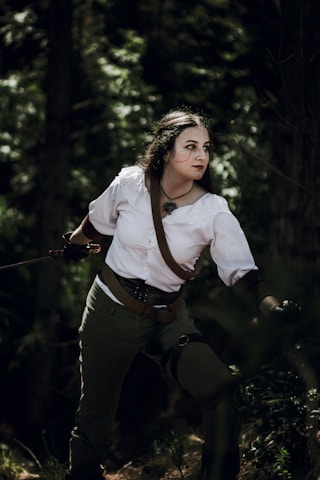 A person with long dark hair and painted face is standing in a forest. They are dressed in historical or fantasy-themed attire, including a white blouse and dark trousers, with a brown belt and strap. They are holding a long sword, prepared for action, and are looking intently to the side.