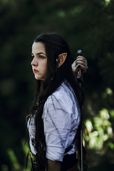 A person with long dark hair and elf-like ears is looking intently to the side. Wearing a white shirt with black detailing, they hold a sword over their shoulder. The background is a blurred green forest environment, giving a mystical vibe.
