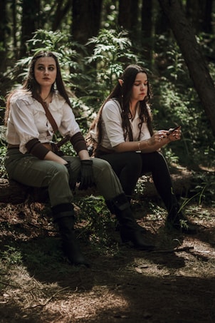 Two individuals dressed in fantasy-style clothing sit on a log in a forest setting. They appear deep in thought, with one holding a small object in their hands. The forest is densely wooded, with filtered sunlight casting shadows around them.