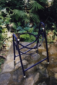 A folded metal chair sits on a stone surface surrounded by lush green foliage, including various types of ferns and leaves. The metal frame is predominantly blue with white-tipped legs, and the scene is illuminated by natural sunlight, creating a peaceful and serene outdoor setting.