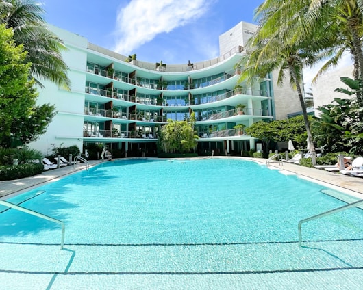 A modern, multi-story hotel or apartment building with curved balconies overlooks a large, turquoise swimming pool. The building is surrounded by lush green palm trees and other tropical vegetation. Sun loungers and umbrellas are arranged around the pool area, creating a relaxing and inviting atmosphere.