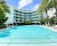 A modern, multi-story hotel or apartment building with curved balconies overlooks a large, turquoise swimming pool. The building is surrounded by lush green palm trees and other tropical vegetation. Sun loungers and umbrellas are arranged around the pool area, creating a relaxing and inviting atmosphere.