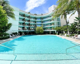 A modern, multi-story hotel or apartment building with curved balconies overlooks a large, turquoise swimming pool. The building is surrounded by lush green palm trees and other tropical vegetation. Sun loungers and umbrellas are arranged around the pool area, creating a relaxing and inviting atmosphere.